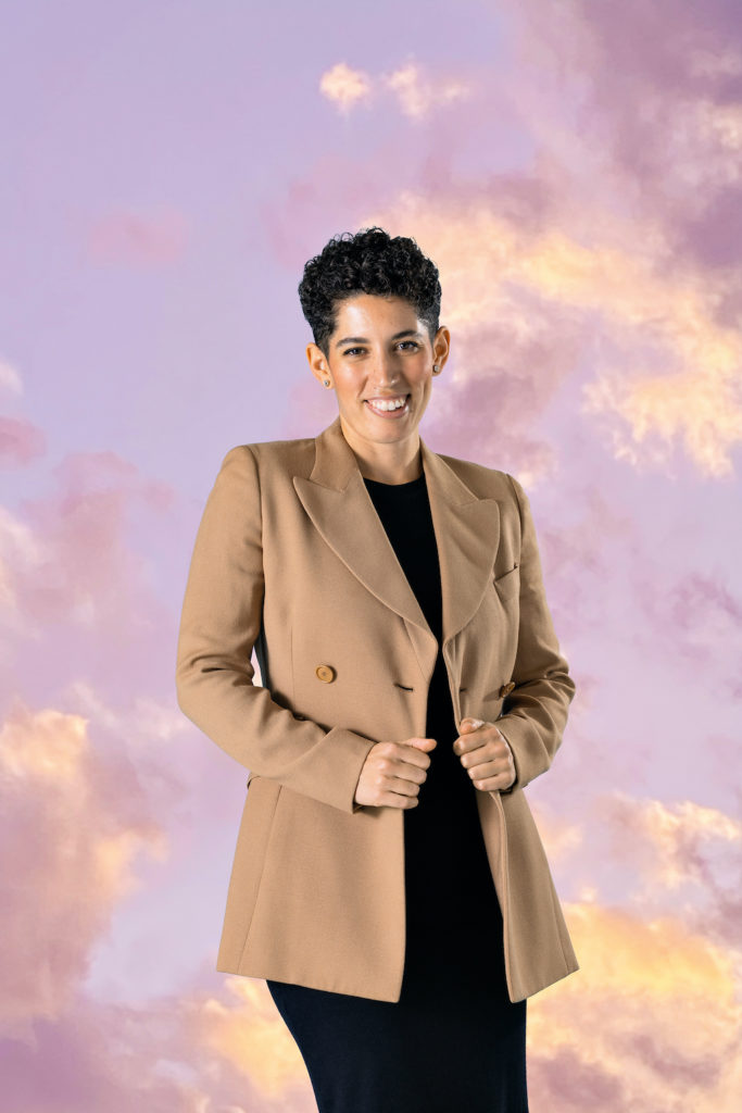 Photo of Aliah El-houni standing in front of a background of a light purple sky with light yellow and deeper purple clouds.