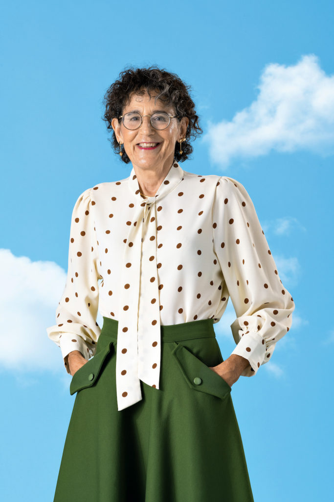 Photo of Ginevra Saylor standing, hands in pockets, in front of a blue sky background with white clouds.