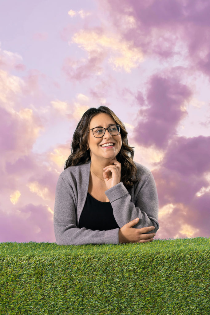 Photo of Leora Smith, her elbows are resting on a ledge of green grass with a background of light pink sky with purple clouds behind her.