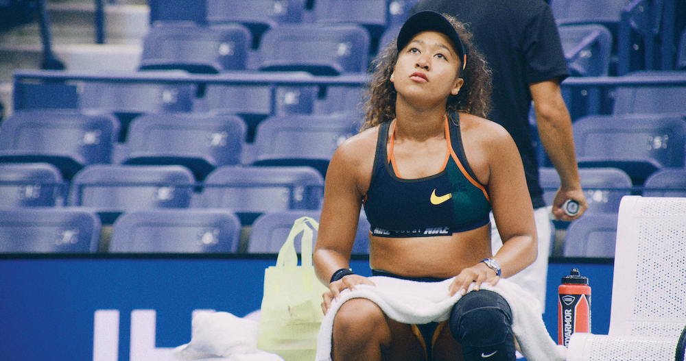 image of Naomi Osaka from the Netflix docuseries. Naomi is sitting on the sidelines of the tennis court looking upwards.