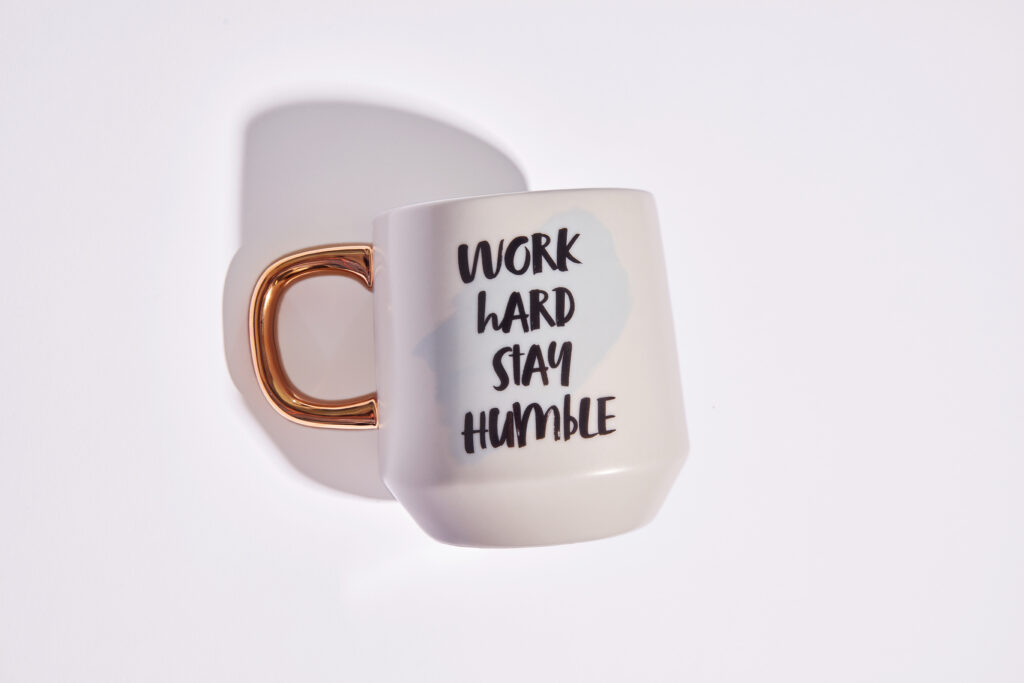 White mug with a gold handle with the words "word hard, stay humble" on it